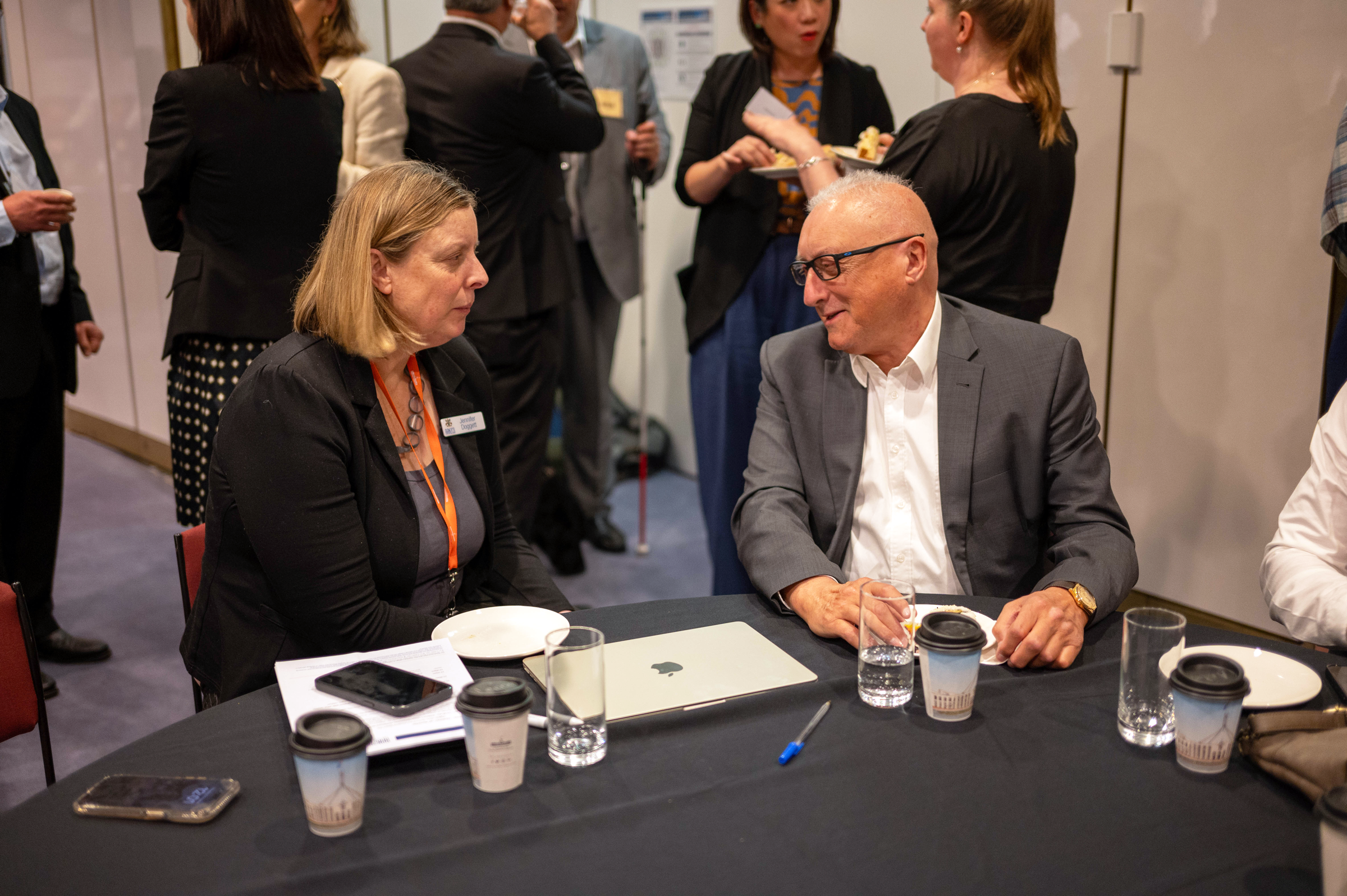Two people in business attire sit at a table in discussion, surrounded by others networking.
